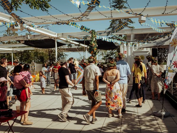 Eine Menschenmenge, viele tragen Blumenkränze auf dem Kopf, versammelt sich unter Girlanden von schwedischen Flaggen bei einer Outdoor-Veranstaltung. Im Hintergrund ist ein IKEA-Logo auf einem Banner zu sehen. Die Szene vermittelt eine festliche Atmosphäre.