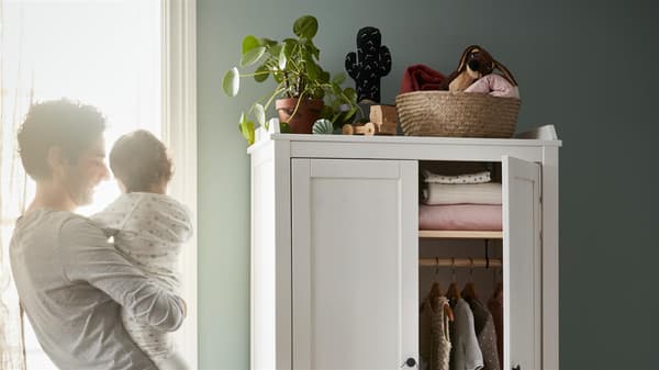 eine Mutter die Ihr Kind in den Armen hällt, neben ihr ein weißer Schrank, darauf sind Kakteen zu sehen.