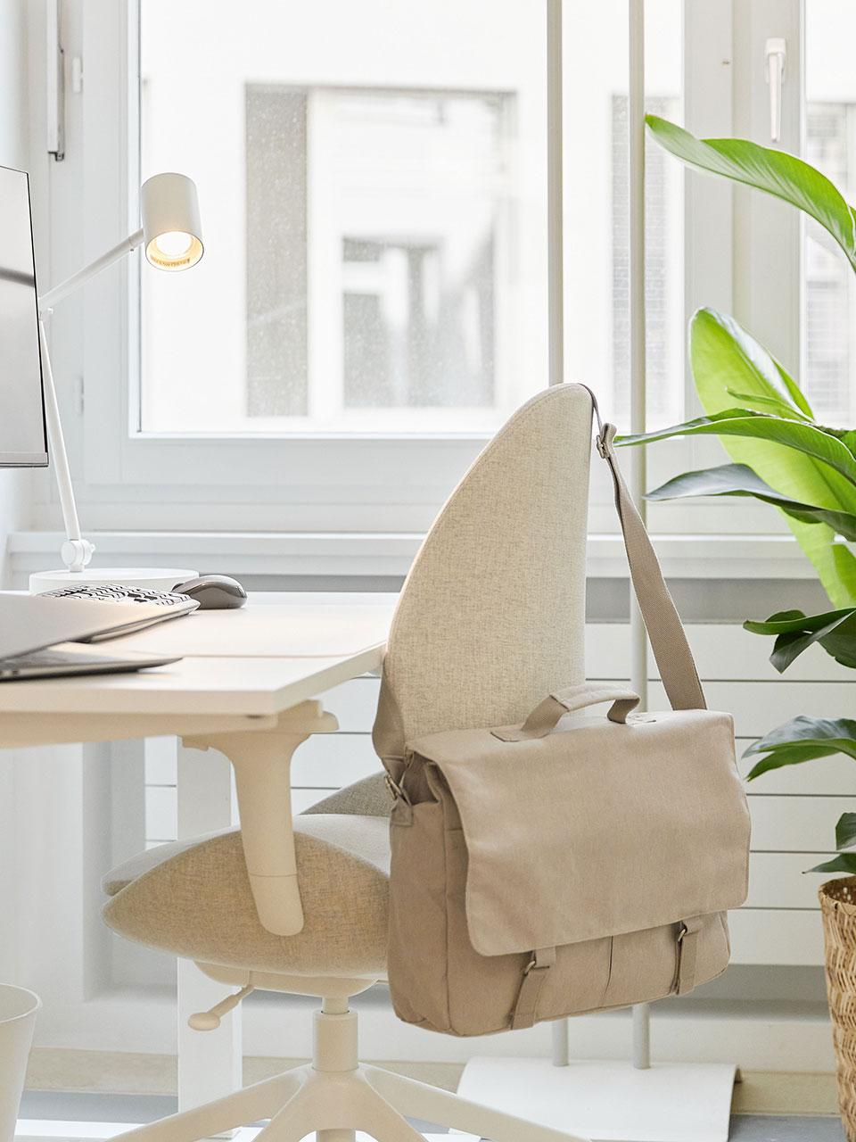 Eine Nahaufnahme eines naturfarbenen Schreibtischstuhls mit Stoffbezug und weissen Armlehnen, an dem eine beige Arbeitstasche hängt. Der Stuhl steht vor einem weissen Schreibtisch an einem Fenster. 