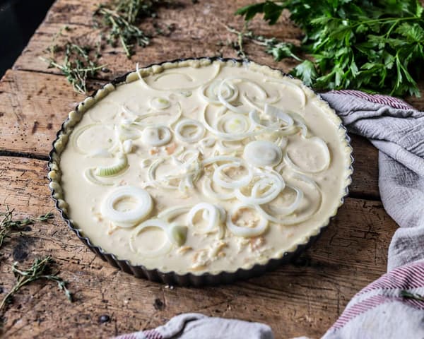 Eine noch ungebackene Midsommar Quiche mit Pilzen steht auf einer Holzplatte.