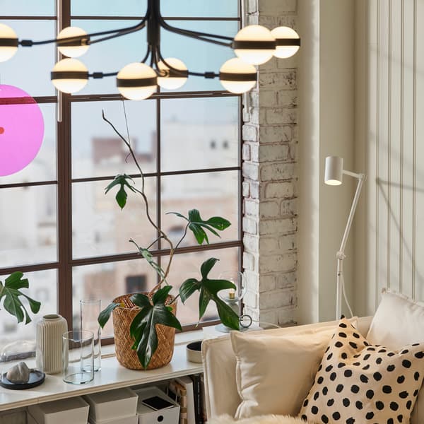 Eine NYMÅNE Stand-/Leseleuchte steht neben einem Sofa an einem Fenster. Von der Decke hängt ein 8-armiger STORSLINGA LED-Kronleuchter herab.