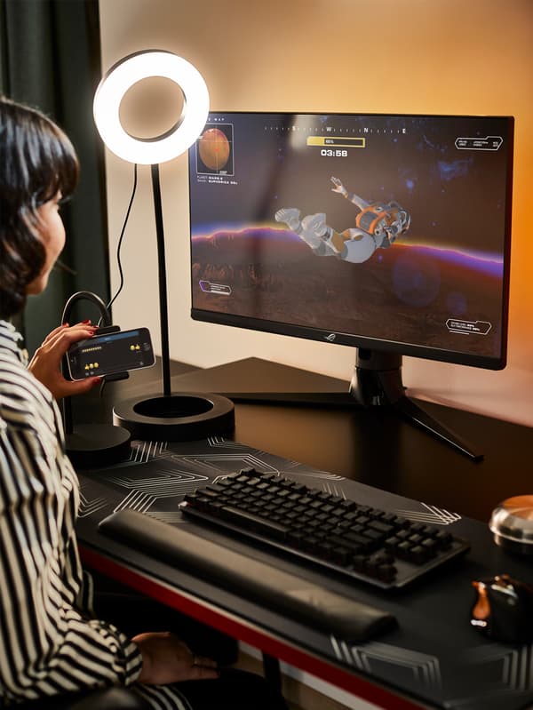 Eine Person, die mit einem Telefon hantiert, sitzt an einem schwarzen HUVUDSPELARE Gamingschreibtisch mit einer eingeschalteten LÅNESPELARE Ringleuchte, einer Tastatur und einem Bildschirm.