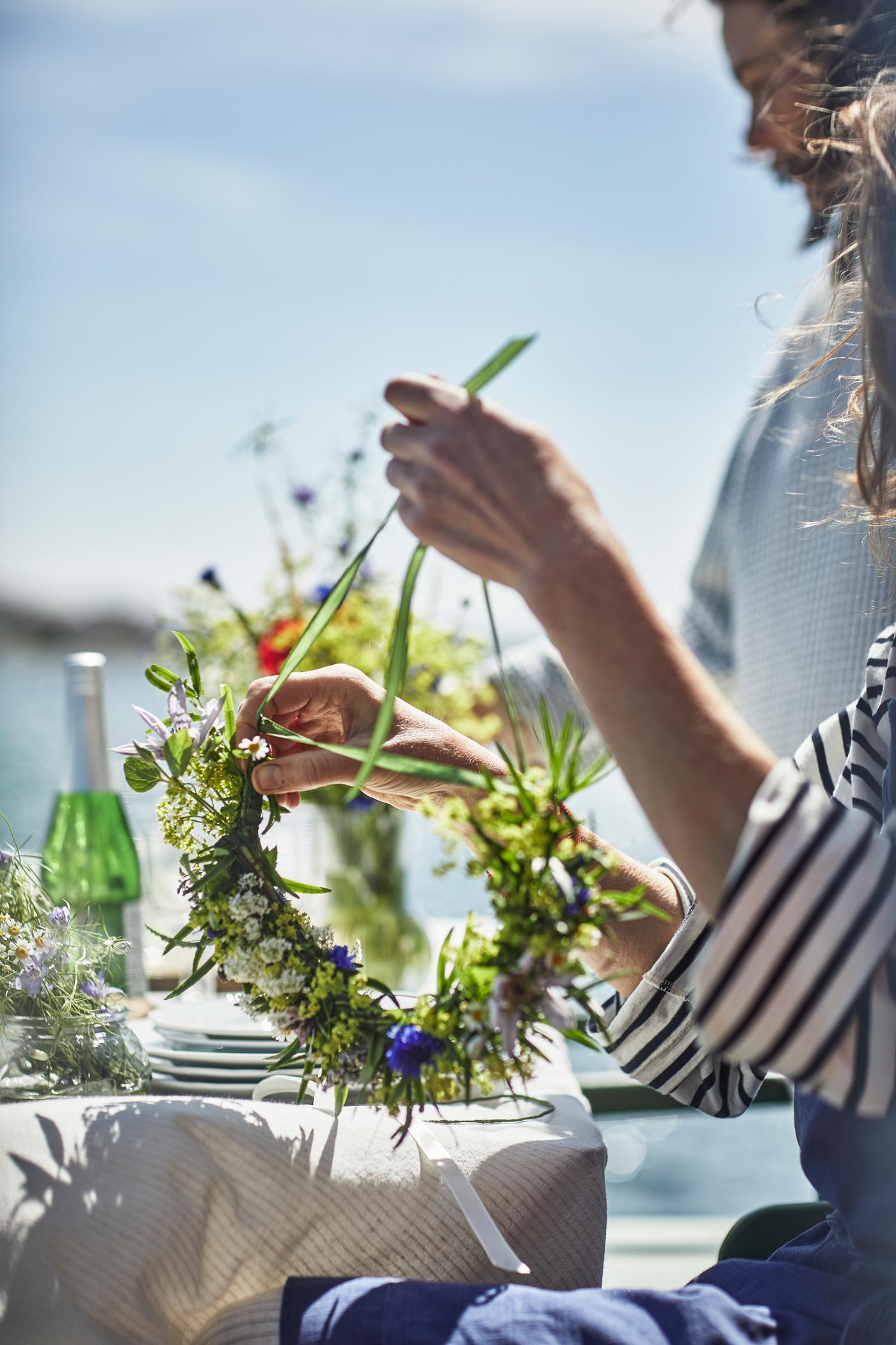 Eine Person gestaltet mit den Händen einen Blumenkranz aus frischen Wiesenblumen an einem gedeckten Tisch im Freien. Im Hintergrund sind Wasser, Geschirr und Glasflaschen im warmen Sonnenlicht zu sehen.