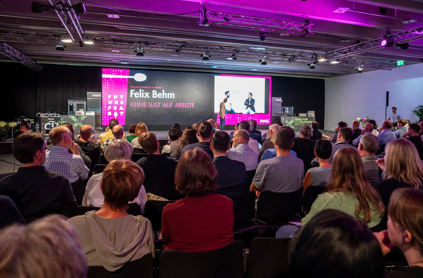 Eine Person hält auf einer Bühne vor Publikum einen Vortrag, während auf einer großen Leinwand eine Präsentation mit dem Titel „Felix Behm – Keine Lust auf Arbeit?“ angezeigt wird.