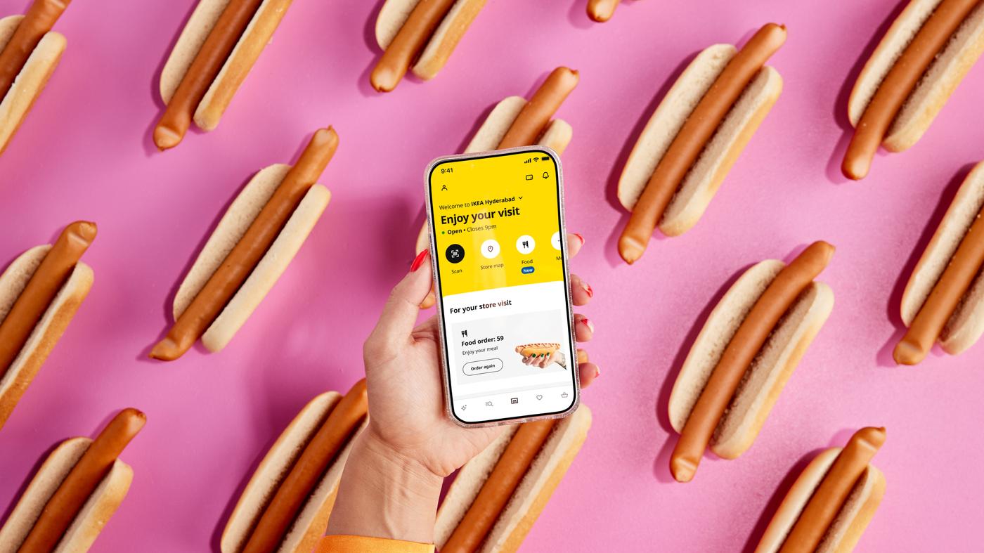 Eine Person hält ein Handy mit einer geööfneten IKEA App und im Hintergrund sind diverse Hot-Dogs zu sehen