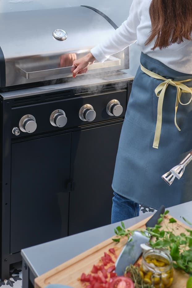 Eine Person hält eine Grillzange in der Hand und ist dabei, den Deckel des Gasgrills einer GRILLSKÄR Außenküche zu öffnen.