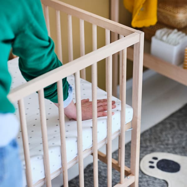 Eine Person in einem grünen Pullover drückt mit der Hand auf die Ecke einer Matratze, die mit einem gepunkteten LENAST Spannbettlaken bezogen ist und in einem SNIGLAR Babybett liegt.