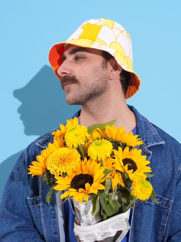 Eine Person in einer Jeansjacke hat einen Hut aus der 3 Meter langen SANDETERNELL Stoffbahn in Gelb auf und hält einen Strauß Sonnenblumen. 
