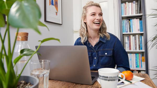 Eine Person lacht, während sie an einem Tisch an ihrem Laptop sitzt. Im Hintergrund ist ein Bücherregal zu sehen.