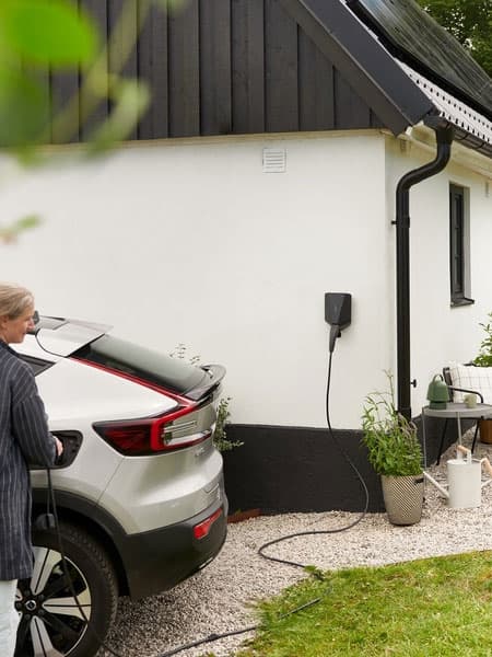 Eine Person lädt ein Elektrofahrzeug an einer Wandladestation vor einem modernen Haus mit Kiesauffahrt.