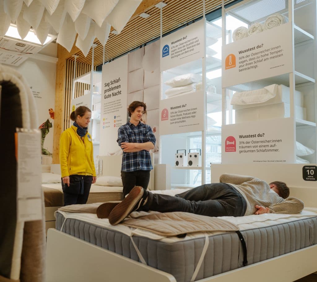 Eine Person liegt in einem Einrichtungshaus auf einem Bett. Die Person davor lässt sich von einer IKEA Mitarbeiterin beraten. 