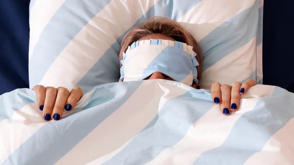 Eine Person liegt mit einer blau-weiß gestreiften Bettdecke bis an die Nase hochgezogen auf einem Bett und hat eine farblich passende Schlafmaske auf.