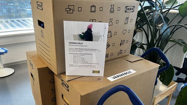 Eine Person mit rosa Mütze sitzt mit dem Rücken zur Kamera in einem hellen Loungesessel; eine dunkelgraue Decke ist über die Schultern gelegt und fällt bis zum Boden, in der Hand eine Tasse mit Heißgetränk. Vor weißem Hintergrund ist rechts eine Lichterkette zu sehen; Jeans und grüne Sneaker der Person sind sichtbar.
