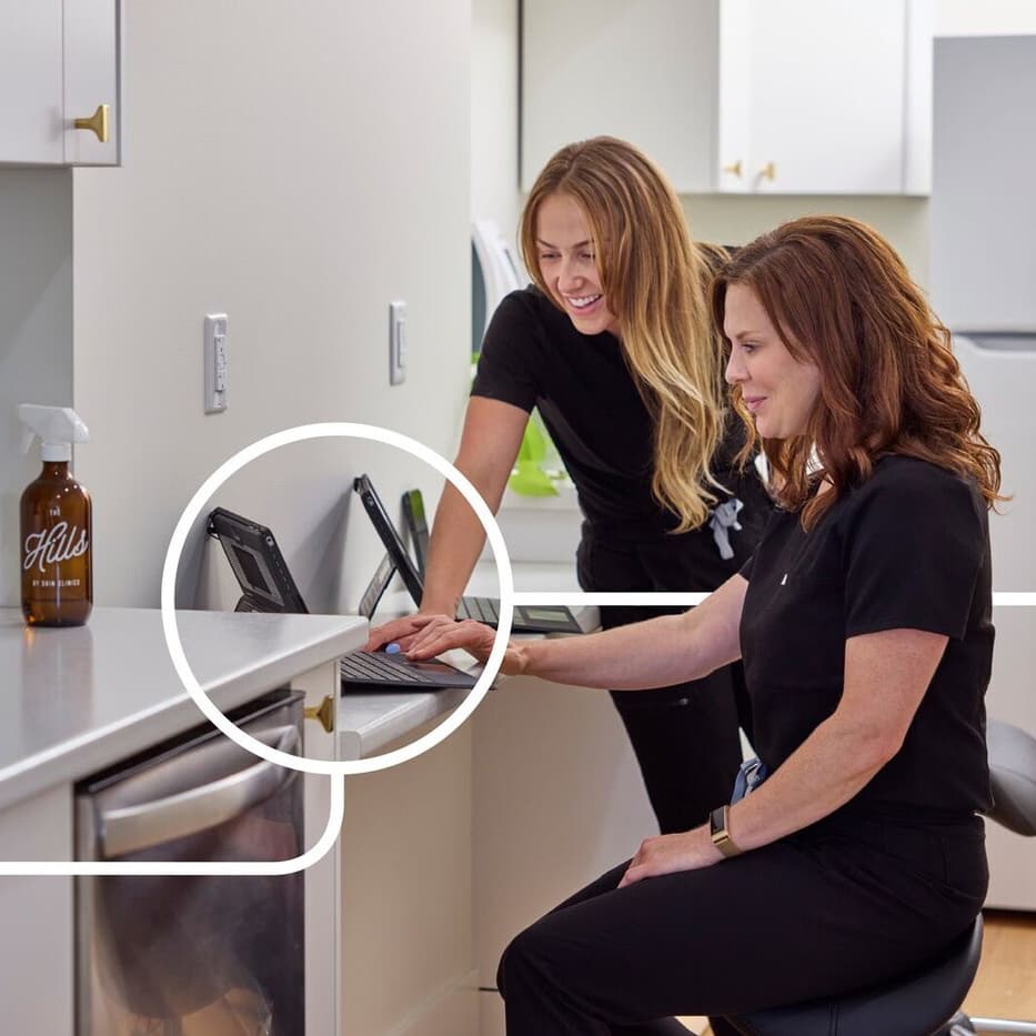 Eine Person sitzt an einem Schreibtisch und bedient einen Laptop. Eine zweite Person schaut ihr über die Schulter und hilft ihr.