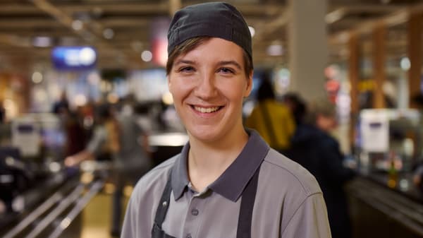 Eine Person steht im Vordergrund und trägt graue Arbeitskleidung.