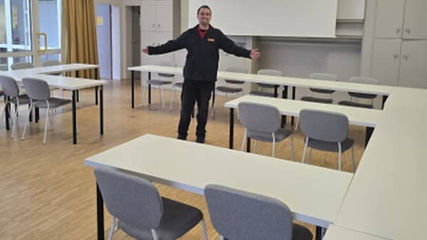 Eine Person steht mit ausgebreiteten Armen in einem hellen Schulungsraum mit mehreren weißen Tischen und grauen Stühlen. Die Tische sind in Reihen angeordnet. Im Hintergrund sind Schränke, eine Projektionsfläche und Fenster zu sehen. Der Raum wirkt neu eingerichtet und für Schulungen vorgesehen.