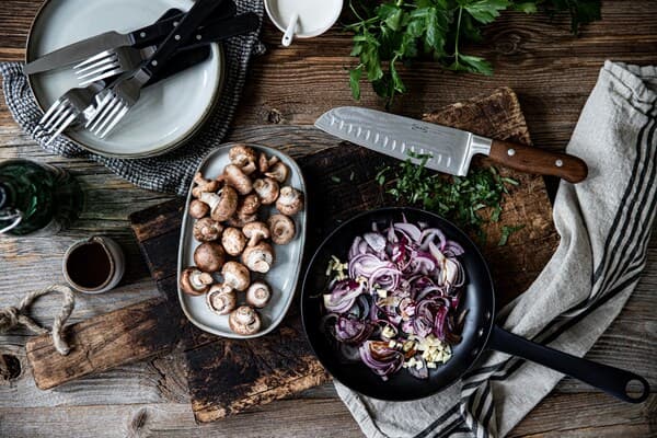 Eine Pfanne mit Zwiebeln und Knoblauch, daneben befindet sich ein Teller mit frischen Pilzen.