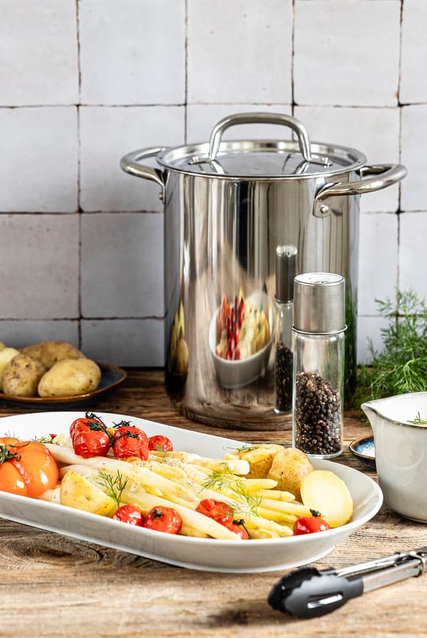 Eine Portion frisch zubereiteter Spargel mit Kartoffeln, geschmorten Tomaten und Zitronen-Dill-Soße steht in einer Küche.
