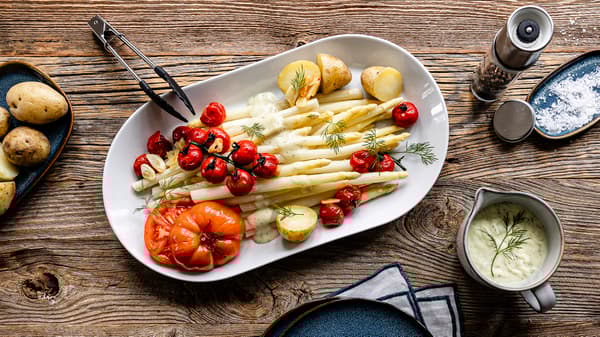 Eine Portion frisch zubereiteter Spargel mit Kartoffeln, geschmorten Tomaten und Zitronen-Dill-Soße steht servierbereit auf einem Holztisch.