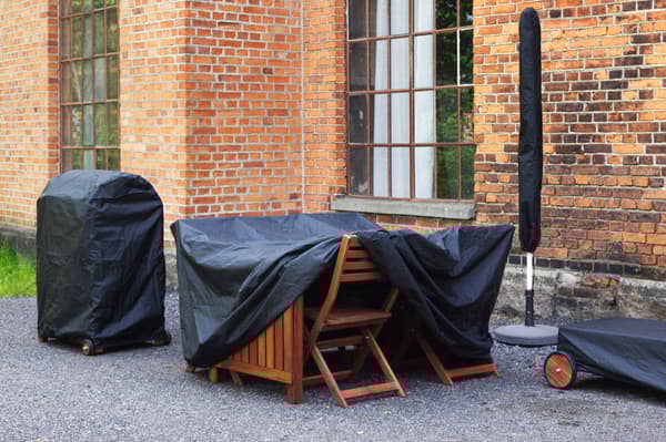Eine schwarze TOSTERÖ Abdeckhaube auf über einem Grill, auf einem Gartentisch mit Stühlen und auf einer Gartenliege.  