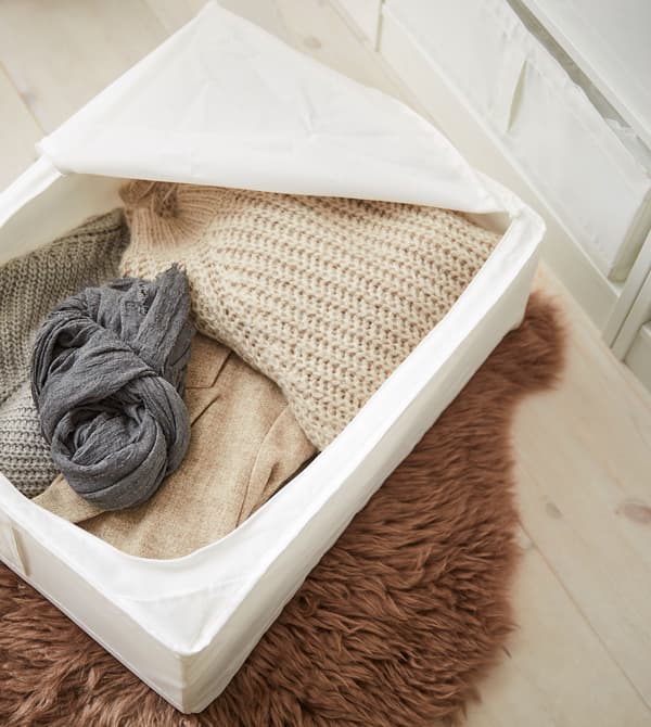 Eine SKUBB Tasche in Weiss gefüllt mit Kleidung in neutralen Farben, auf einem flauschig wirkenden, braunen Teppich.