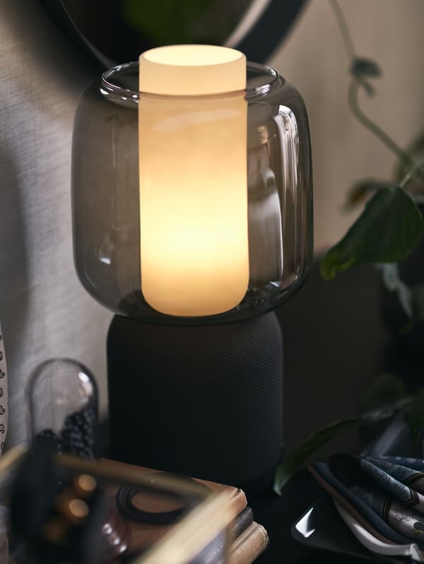 Eine SYMFONISK WiFi-Leuchte mit Speaker in Schwarz mit einem Leuchtenschirm aus Glas hängt an einer grauen Wand. U. a. ist eine Pflanze zu sehen.