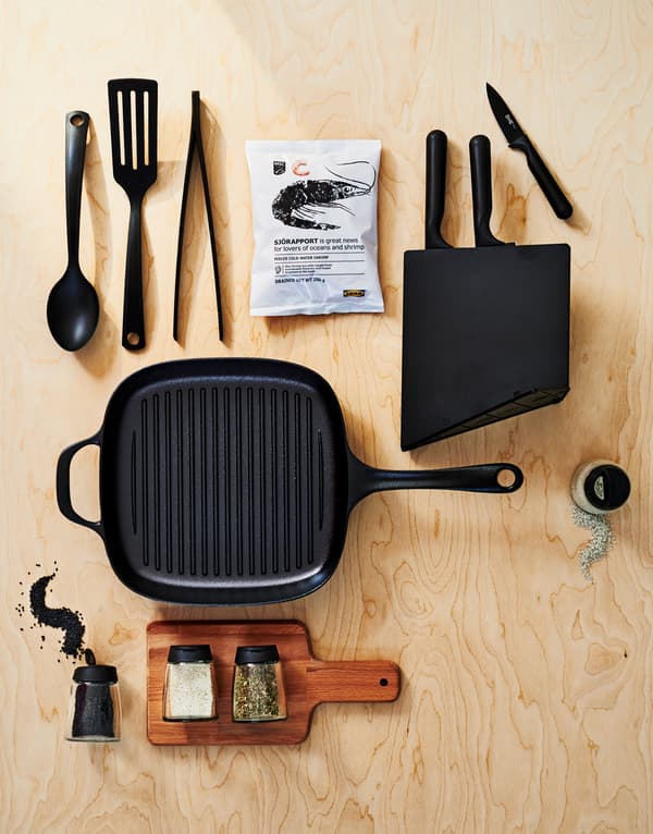 Eine VARDAGEN Grillpfanne aus Gusseisen mit den GNARP Küchenutensilien, einem Messerblock, einem Schneidebrett und Gewürzen auf einer Oberfläche aus Holz.