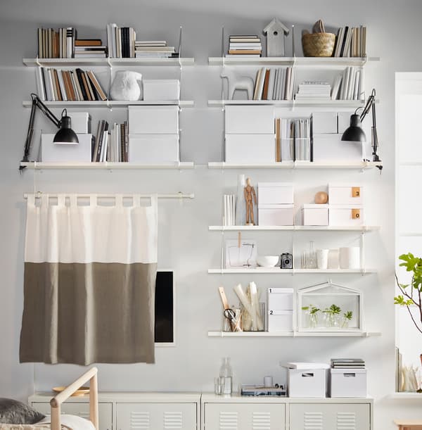 Eine weiss gestrichene Wand mit Regalen, u. a. mit TJENA Kästen mit Deckeln. Die Anordnung ist sehr symmetrisch.
