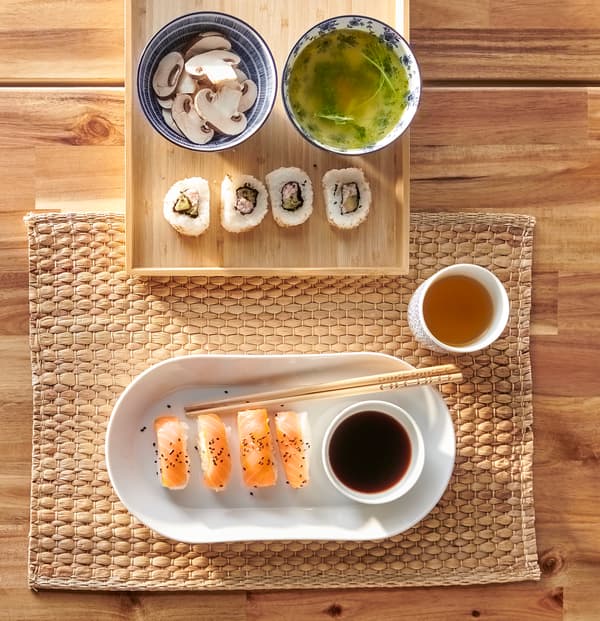 Eine weisse IKEA 365+ Servierplatte mit Sushi und weissen und blauen ENTUSIASM Schüsseln aus Keramik mit Suppe und Pilzen auf einem Bambustablett und einem Tischset.