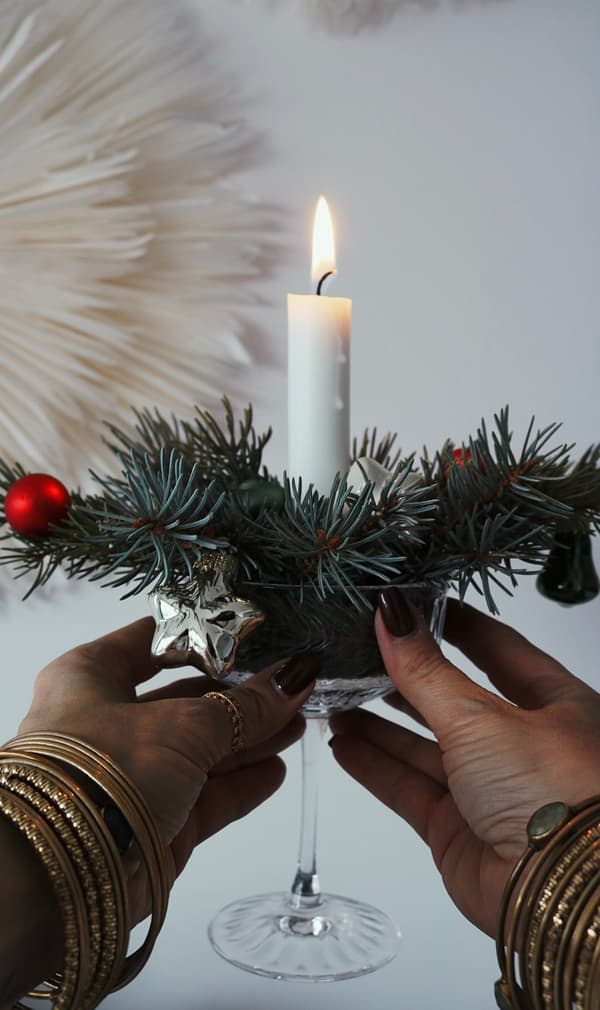 Eine weisse Kerze in einem Glashalter ist mit Tannenzweigen und roten Kugeln geschmückt. Hände mit Armreifen halten sie und vermitteln eine festliche, elegante Atmosphäre.