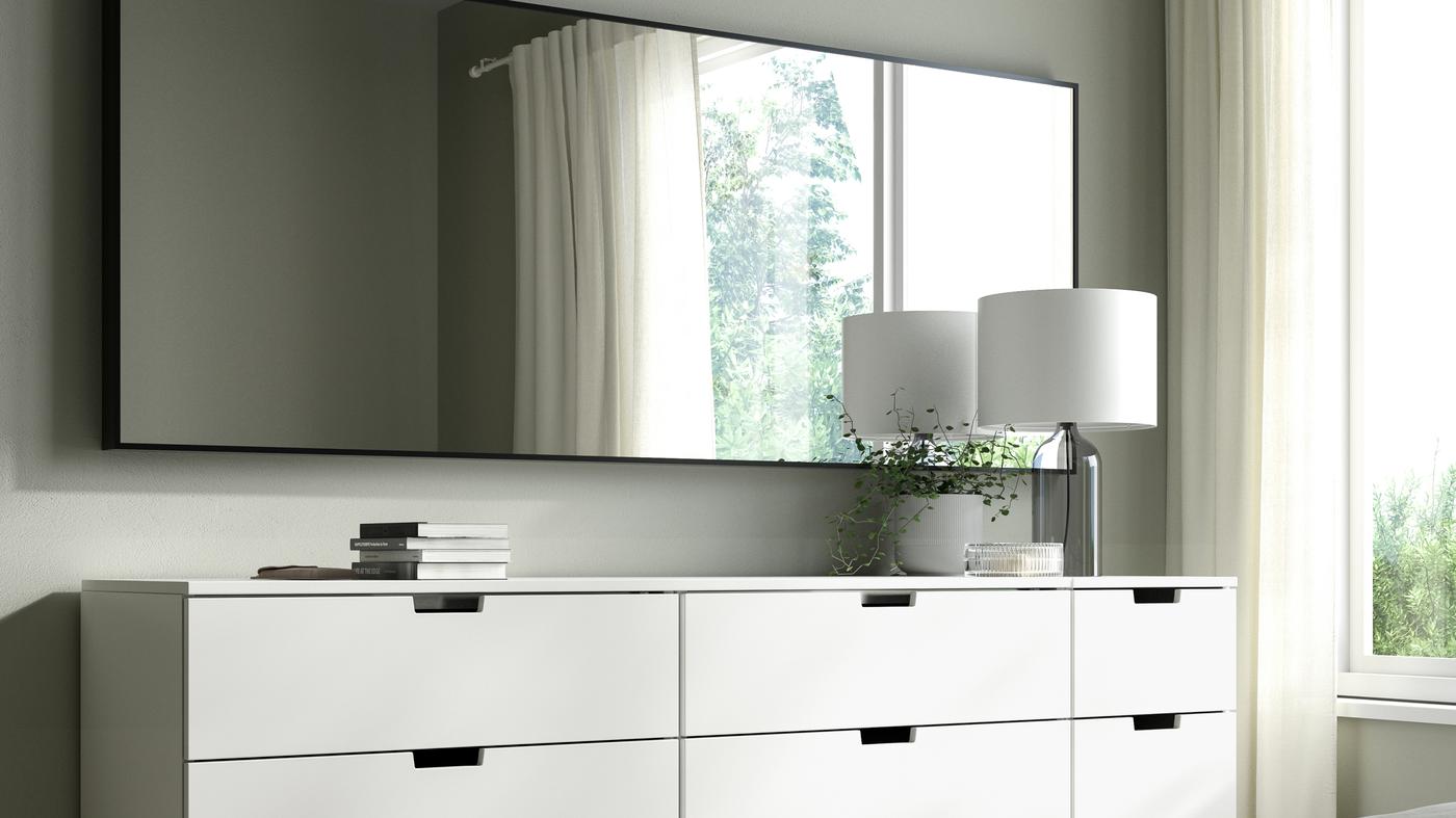 Eine weiße Kommode mit Dekoelemten wie Büchern und einer Lampe, so wie einem großen Spiegel quer an der Wand.