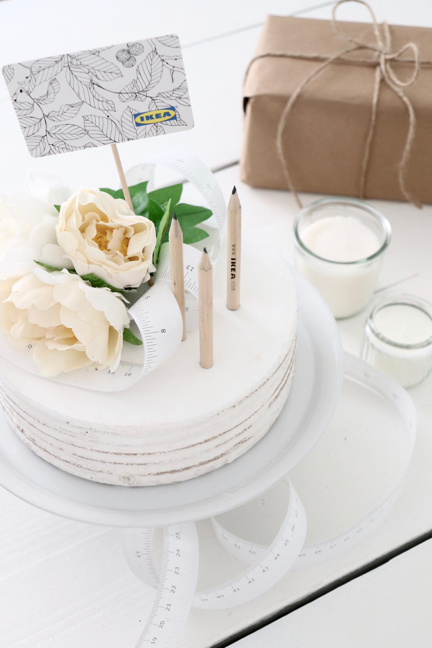 Eine weiße Torte mit Blumendeko und IKEA Bleistiften sowie IKEA Geschenkkarte von oben.