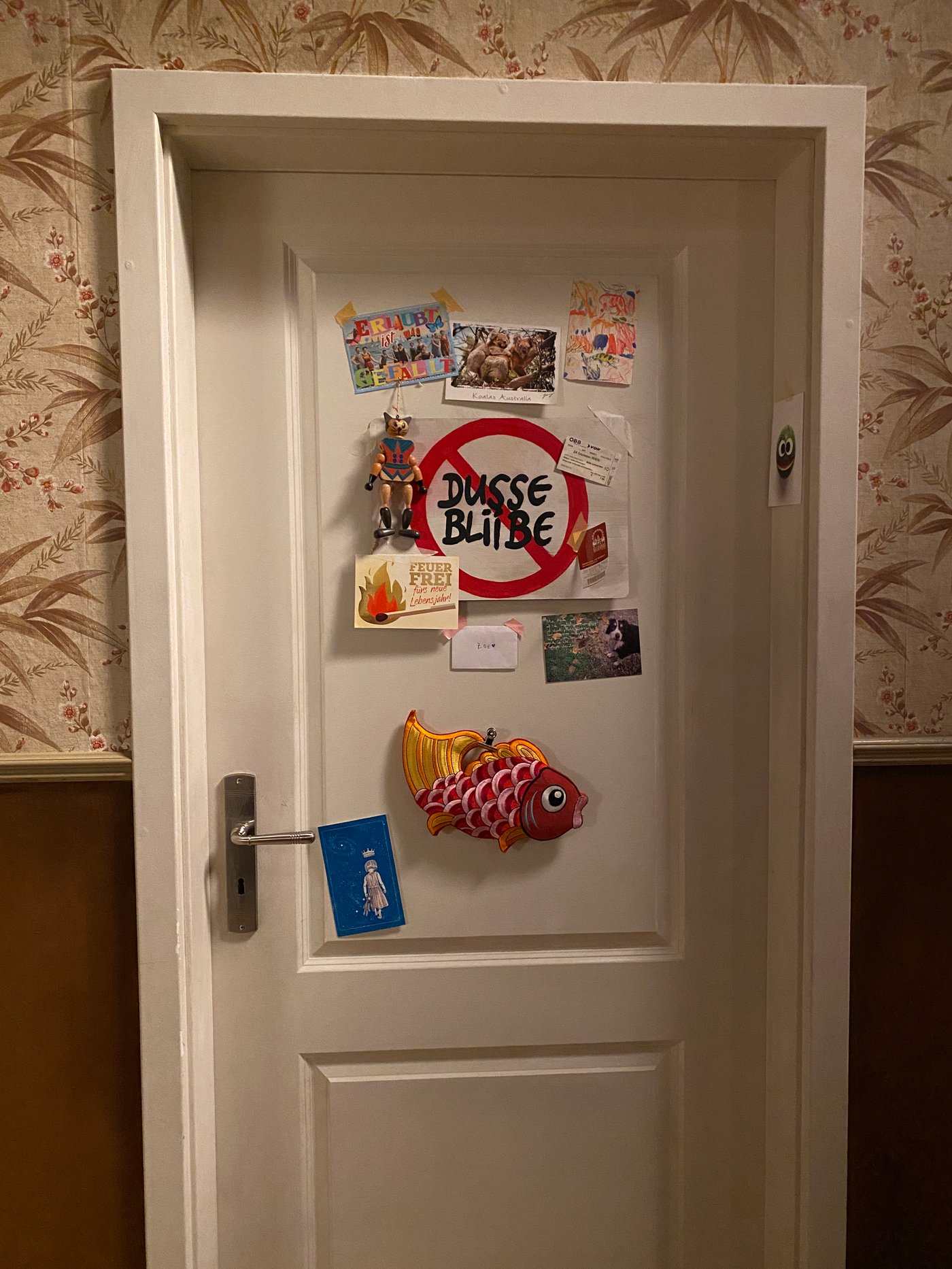 Eine weisse Zimmertür mit verschiedenen Postkarten und einem Schild mit der Aufschrift: "Dusse Bliiebe"