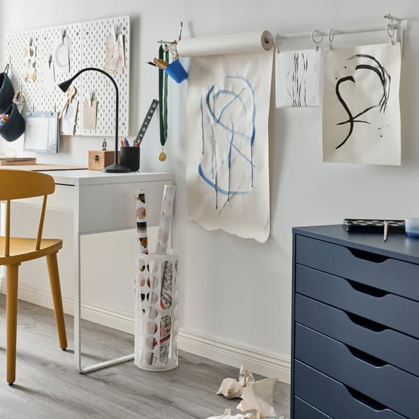Eine Zeichenpapierrolle und Papierblätter mit Zeichnungen darauf hängen an einer an der Wand angebrachten Gardinenstange. Im Vordergrund steht ein blaues Schubladenelement.