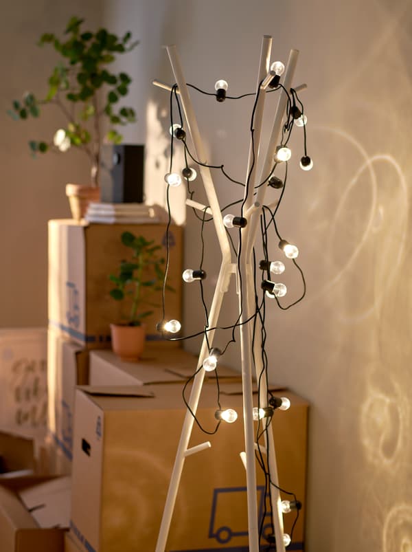 ที่แขวนหมวกและเสื้อโค้ท EKRAR/เอียแครร์ ถูกพันด้วยไฟประดับ LED BLÖTSNÖ/เบลิตสเนอ ที่เปิดอยู่ ถัดจากลังสำหรับขนของที่วางซ้อนกัน
