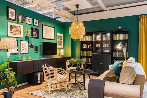 Ekspozycja. Salon z ciemnozielonymi ścianami. Na jednej z nich mnóstwo ramek i jeden telewizor. Na środku pokoju rattanowe fotele przy stoliku, obok sofa.