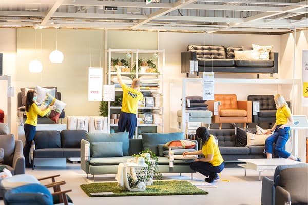 Ekspozycja sklepowa z sofami. Pośród mebli pracownicy w żółtych koszulkach z napisem HEJ! dopracowują ostatnie szczegóły - układają poduszki, dostawiają rośliny.