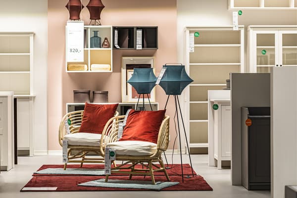 Ekspozycja z dwoma rattanowymi fotelami - jeden stoi za drugim. Na nich czerwone i białe poduszki. Koło nich lampy, a na podłodze białe i czerwone dywany.