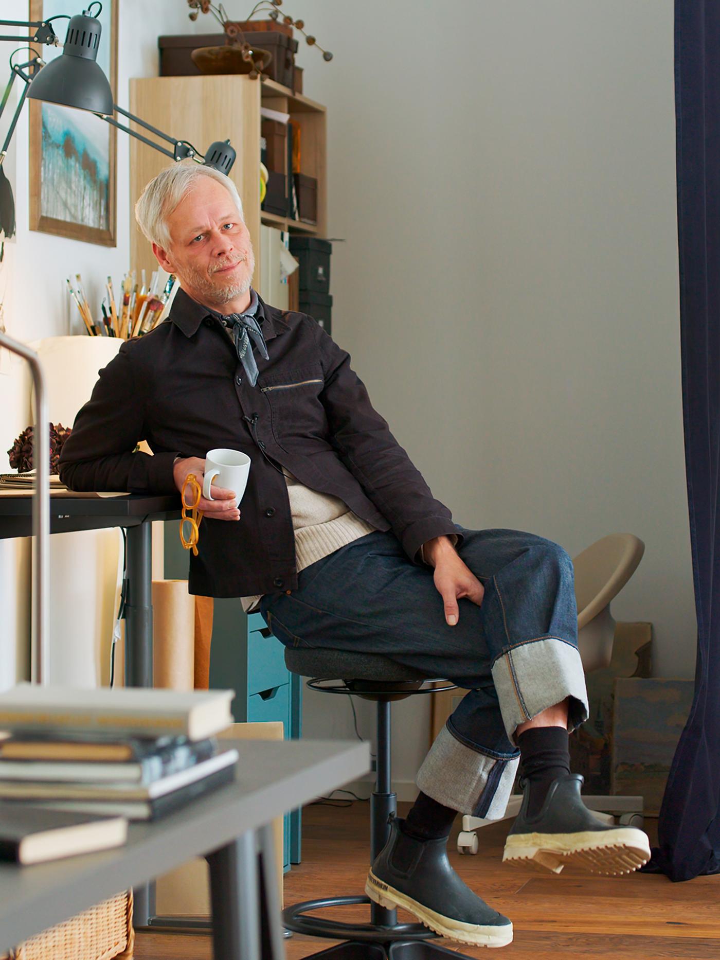 El diseñador Hans Blomquist sentado en un taburete en una habitación luminosa con una taza en la mano.