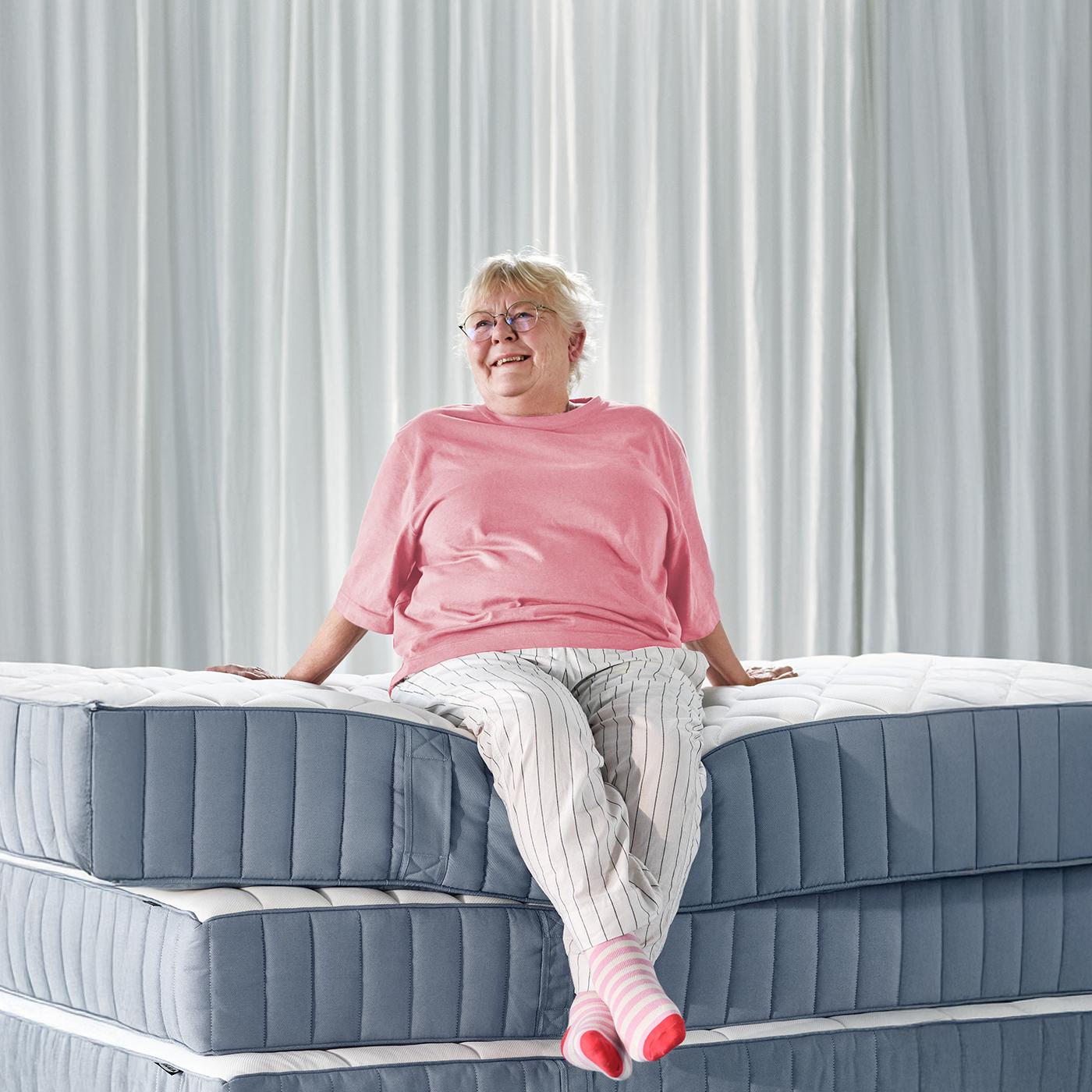 Elderly woman sitting comfortably on a stacked mattress, smiling in a bright modern bedroom