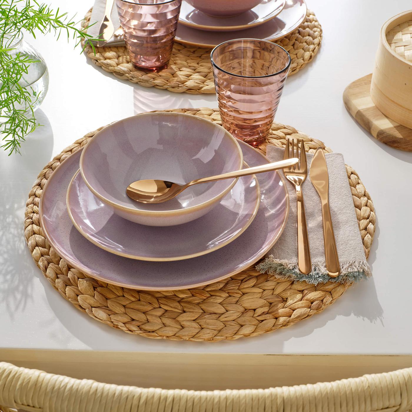 Elegant table setting with stacked purple plates, gold cutlery, pink glass, and bowl on woven placemat with white table surface.