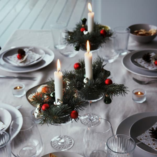 Élégante table de fête avec des bougies allumées sur des couronnes de pin, des assiettes blanches et des verres transparents. Ambiance festive et chaleureuse avec une décoration rouge et verte.