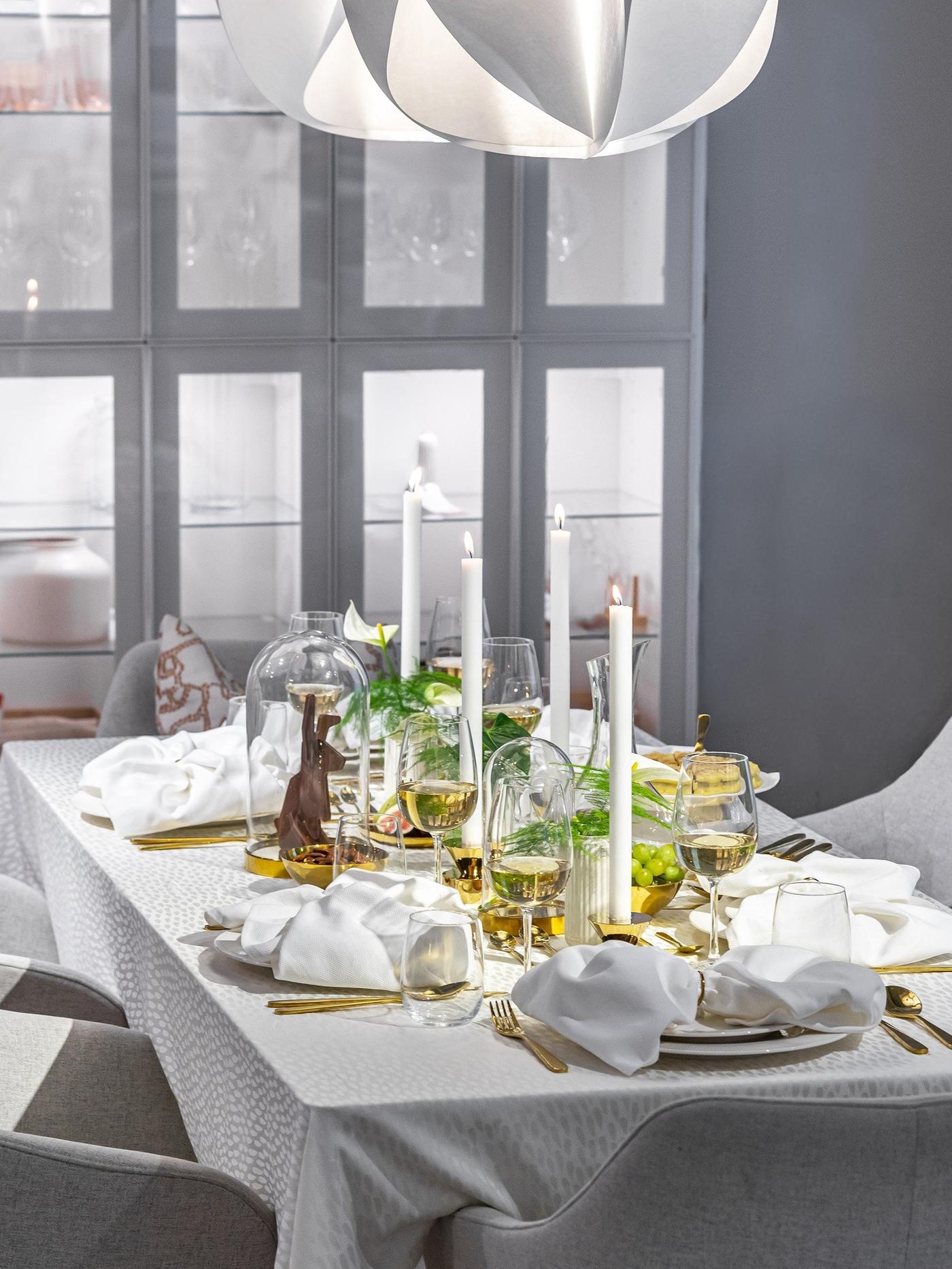 Elegantiškai padengtas valgomojo stalas su balta staltiese, skaidraus stiklo taurėmis, auksinės spalvos įrankiais ir aukštomis žvakėmis.