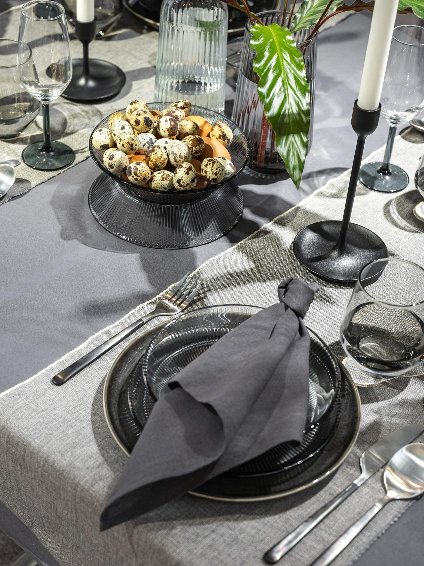 Elegantiško stalo fragmentas su pilkų atspalvių tekstile, moderniais indais, putpelių kiaušiniais ir subtiliai žaliomis šakelėmis.