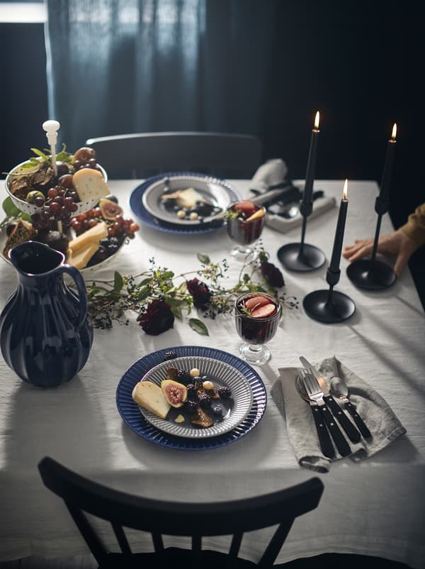Elegantly set table, including a VANLIGEN jug and LIVNÄRA cutlery, on a white tablecloth in an otherwise dark-coloured room.
