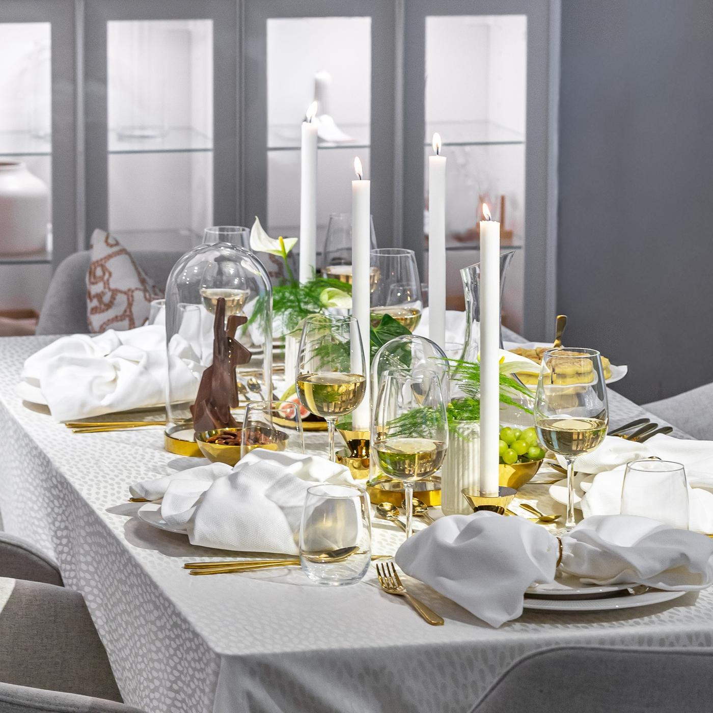 Элегантно накрытый стол с мягкой скатертью в нейтральных тонах, прозрачной посудой, золотистыми столовыми приборами и высокими свечами.