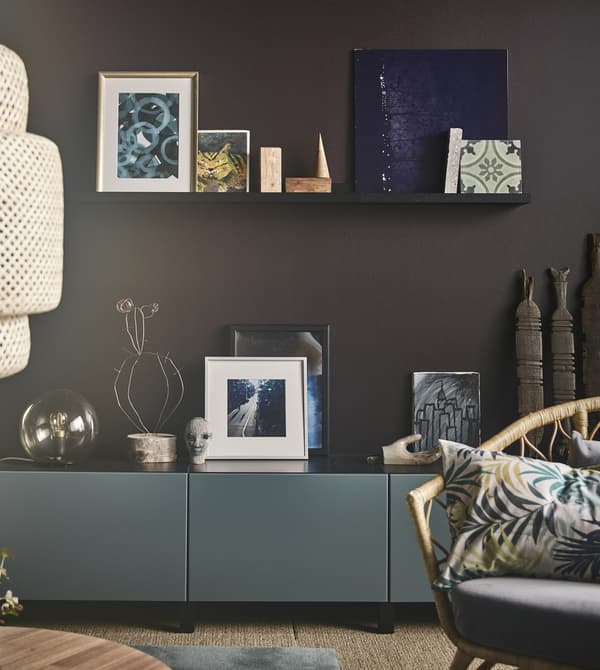 Elementos artísticos colocados en un estante para cuadros en negro en una pared marrón sobre un armario gris-turquesa en un salón moderno.