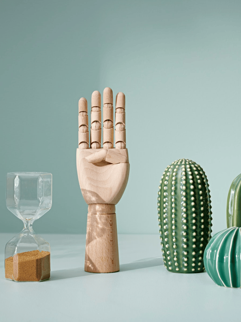 Elementos decorativos contra una pared verde, como una estatua de cactus, un reloj de arena y una mano articulada de madera.