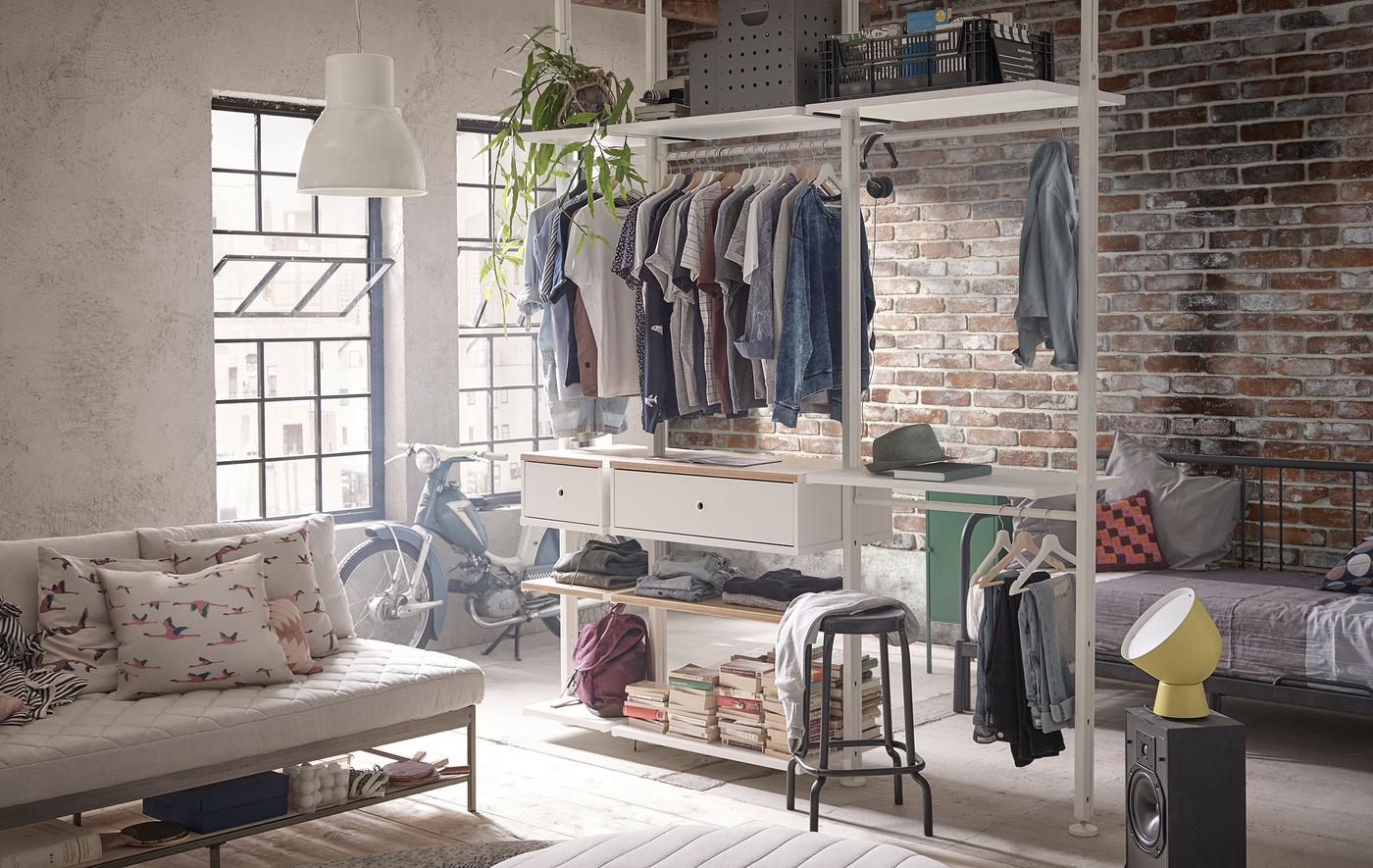 차분한 톤과 벽돌 벽으로 꾸민 로프트 아파트에 옷과 책이 정리되어 있는 ELVARLI 엘발리 수납시스템이 있습니다.