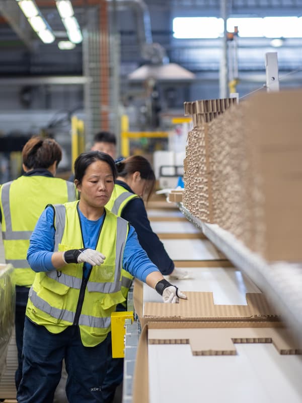 Empat pekerja Asia yang memakai ves keterlihatan tinggi sedang membungkus produk di barisan pemasangan kilang pembekal IKEA.